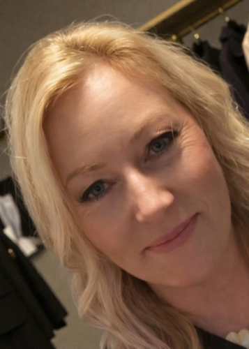 Egy kék szemű, mosolygó szőke nő közeli portréja. A háttér homályos, ruhák és lámpák kivehetők. Női portré, mosoly, szépség, nő, arckép, üzleti portré, önbizalom, elegancia, szőke haj.